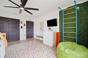 A room with a ceiling fan, a green wall with a climbing wall, a bed, a desk, and a TV.