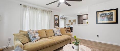 A living room with a sofa, a round coffee table, a ceiling fan, and a kitchen in the background.
