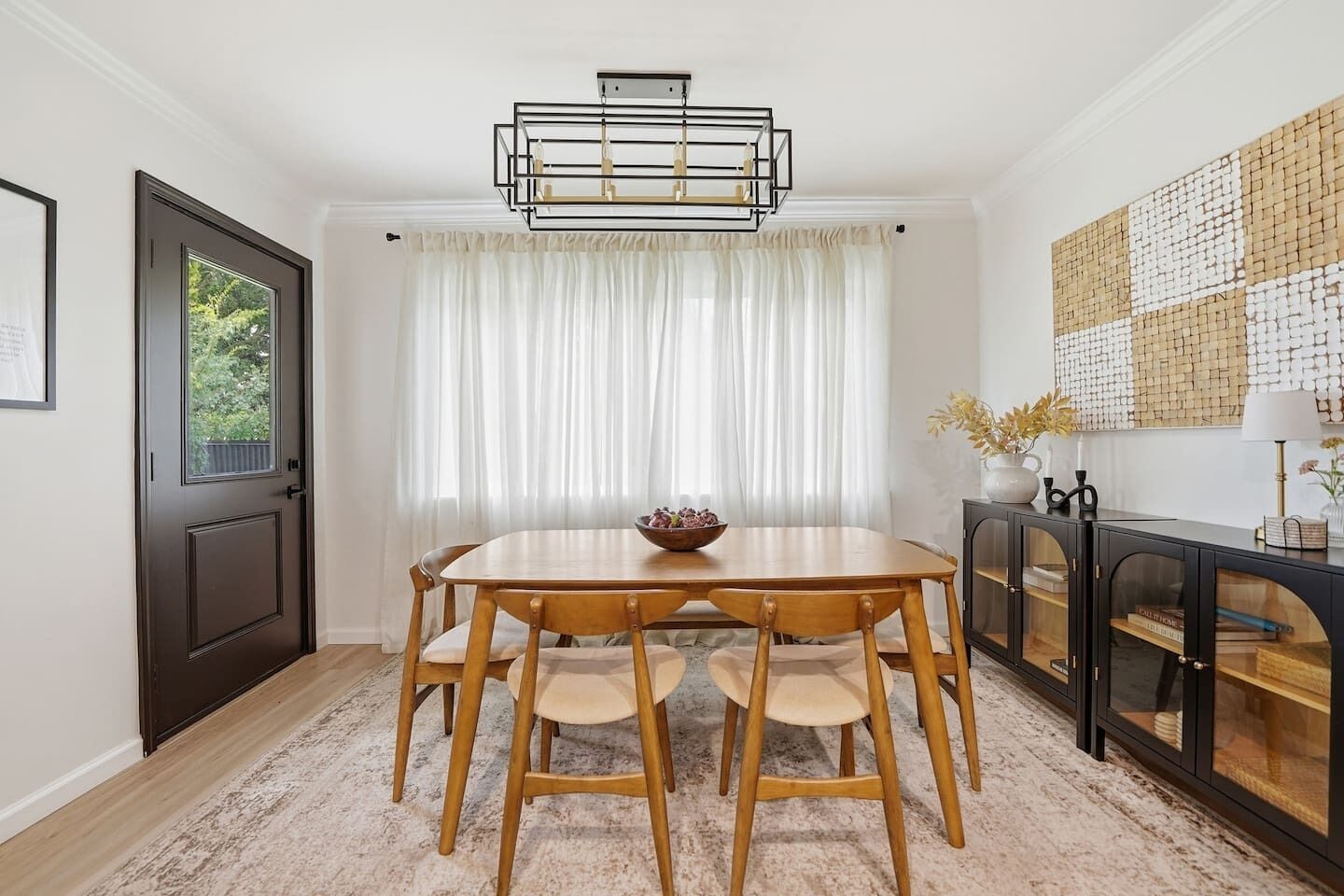 A dining area with a wooden table and chairs, a chandelier, a window with curtains, and a decorative wall piece.