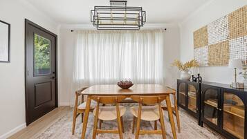A dining area with a wooden table and chairs, a chandelier, a window with curtains, and a decorative wall piece.