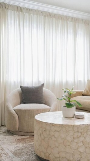 A living room with a round marble coffee table, a beige armchair, a potted plant, and a large window with sheer curtains.