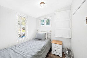 Un dormitorio con una cama individual, una mesita de noche, un ventilador de pared y un pequeño ventanal sobre la cama.