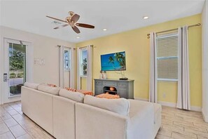 A living room with a beige sofa, a ceiling fan, a television on a stand, and a window with blinds. - Margaritaville 4BR | Sleeps 10 | Near Disney (Kissimmee)