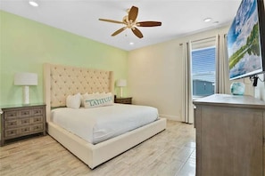 A bedroom with a tufted headboard, a bed with white bedding, a nightstand with a lamp, a ceiling fan, a window with curtains, and a television on a stand. - Margaritaville 4BR | Sleeps 10 | Near Disney (Kissimmee)