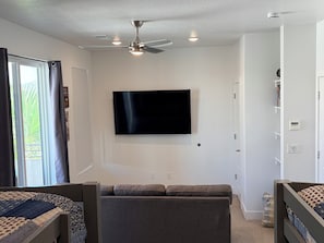 A room with a ceiling fan, a flat-screen TV mounted on the wall, a gray sofa, and a window with curtains. - The Desert Belle, No. 1 Loft at The Lofts,  4BR 3.5BA Southern Utah Retreat (Saint George)