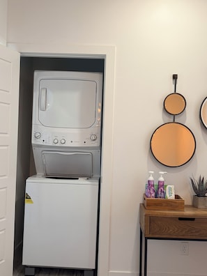A compact laundry room with a stacked washer and dryer, a wooden side table with bottles, and wall-mounted mirrors.