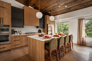 A modern kitchen with wooden cabinets, a central island, bar stools, and a view of greenery outside. - Blue Jay Falls Overlook: Luxury Cabin, Hot Tub, Sauna, Game Room & Views (Ellijay)