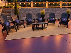 Outdoor seating with Adirondack chairs and a small table, set against a stone wall with string lights. - Blue Jay Falls Overlook: Luxury Cabin, Hot Tub, Sauna, Game Room & Views (Ellijay)