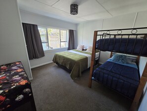 A room with bunk beds, a single bed, a window with curtains, a floral table, and a ceiling light fixture. - Middleton Near The Beach (Paraparaumu Beach)