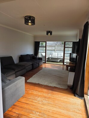 A living room with a sectional sofa, a white rug, wooden flooring, and a view through large windows to an outdoor area with a fence. - Middleton Near The Beach (Paraparaumu Beach)