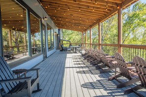 Eine überdachte Terrasse im Freien mit Holzsesseln und -tischen, großen Fenstern und Blick auf Bäume.
