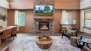 Olohuoneessa on kivinen takka, jonka yläpuolella on seinälle asennettu televisio, ruokailutila, jossa on pöytä ja tuolit, sekä suuri ikkuna, josta avautuu näkymä ulos.
