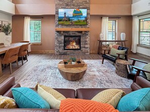 A living room with a stone fireplace, a television mounted above it, a dining area with a table and chairs, and a large window providing a view of the outdoors. - Luxury 4BR Cabin | Lazy River | Near Big Cedar (Hollister)