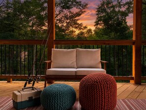 Eine Holzterrasse mit einer Couch und zwei gestrickten Hockern, eine Angel und eine Kühlbox.