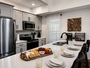 A modern kitchen with stainless steel appliances, a central island with a fruit platter, and a dining area set for four. - Luxury 4BR Cabin | Lazy River | Near Big Cedar (Hollister)