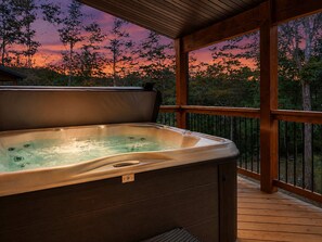 A hot tub on a wooden deck with a view of trees and a sunset.