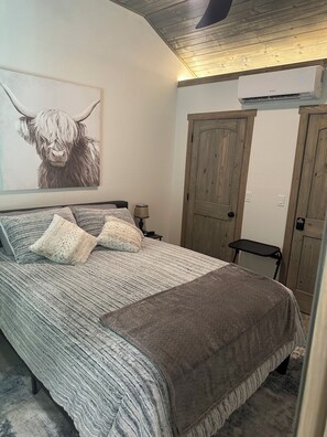 A bedroom with a bed, pillows, a wall-mounted air conditioner, and a wooden door.
