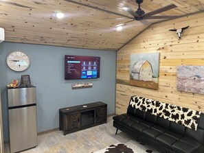 A modern living room with a flat-screen TV, a clock, a refrigerator, a wooden ceiling, and a cow-patterned sofa.