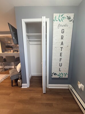 A room with a grey wall, a wooden floor, a white door, a white-framed mirror, a flat-screen TV, a grey sofa, a small table with a vase, and a decorative sign with the word 'GRATEFUL'.