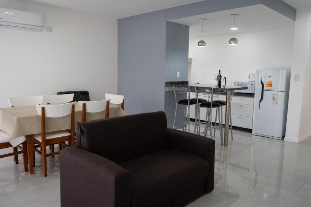 Una sala de estar moderna con sofá, mesa de comedor, sillas y una cocina con refrigerador, estufa y taburetes de bar.
