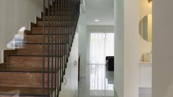 Un escalier avec des marches en bois et des rampes en métal mène à une pièce lumineuse, dotée d’un grand miroir et d’un mur blanc.