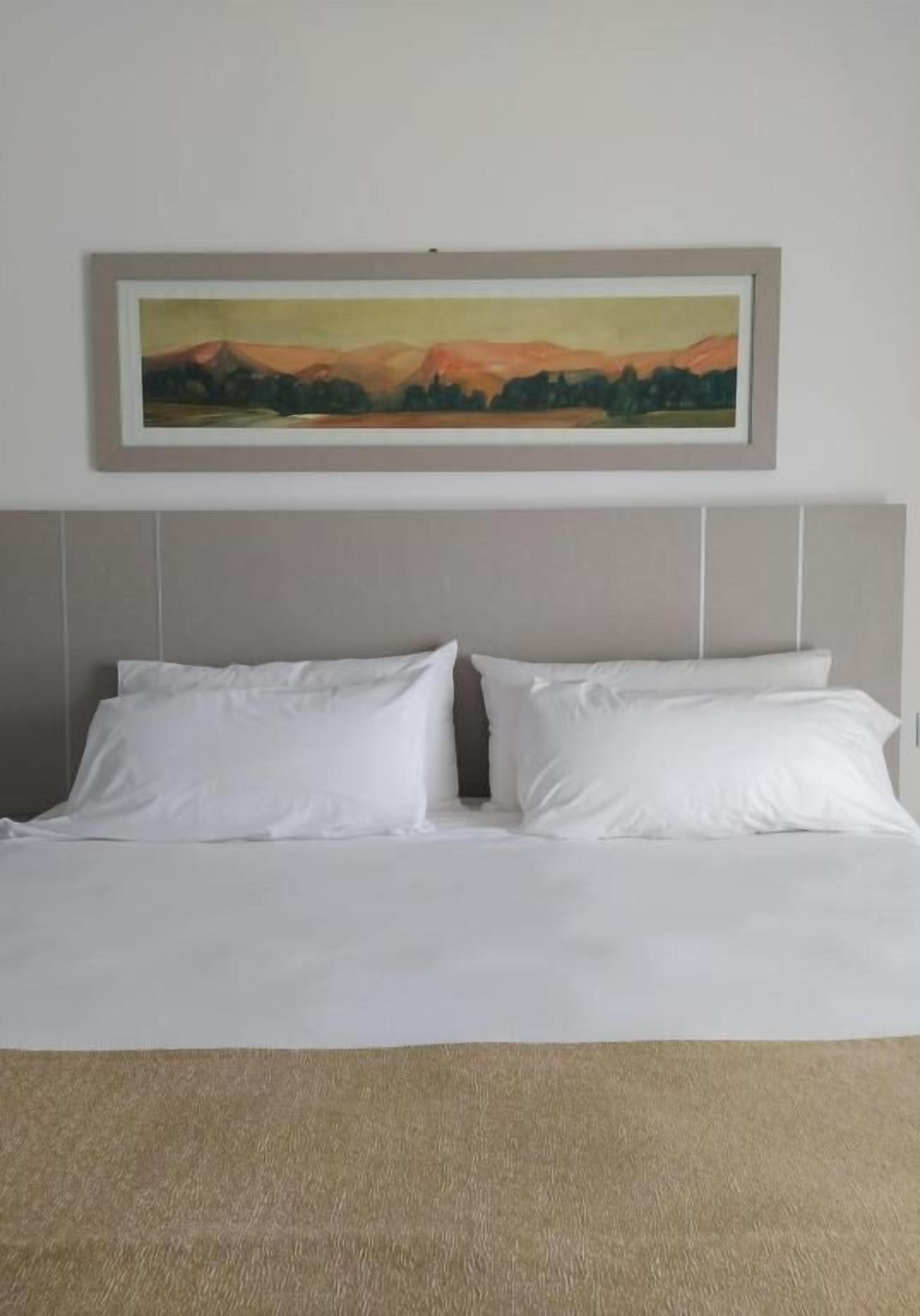 Una cama con ropa de cama blanca y un cabecero, con un cuadro de un paisaje enmarcado encima, y una alfombra color beige.