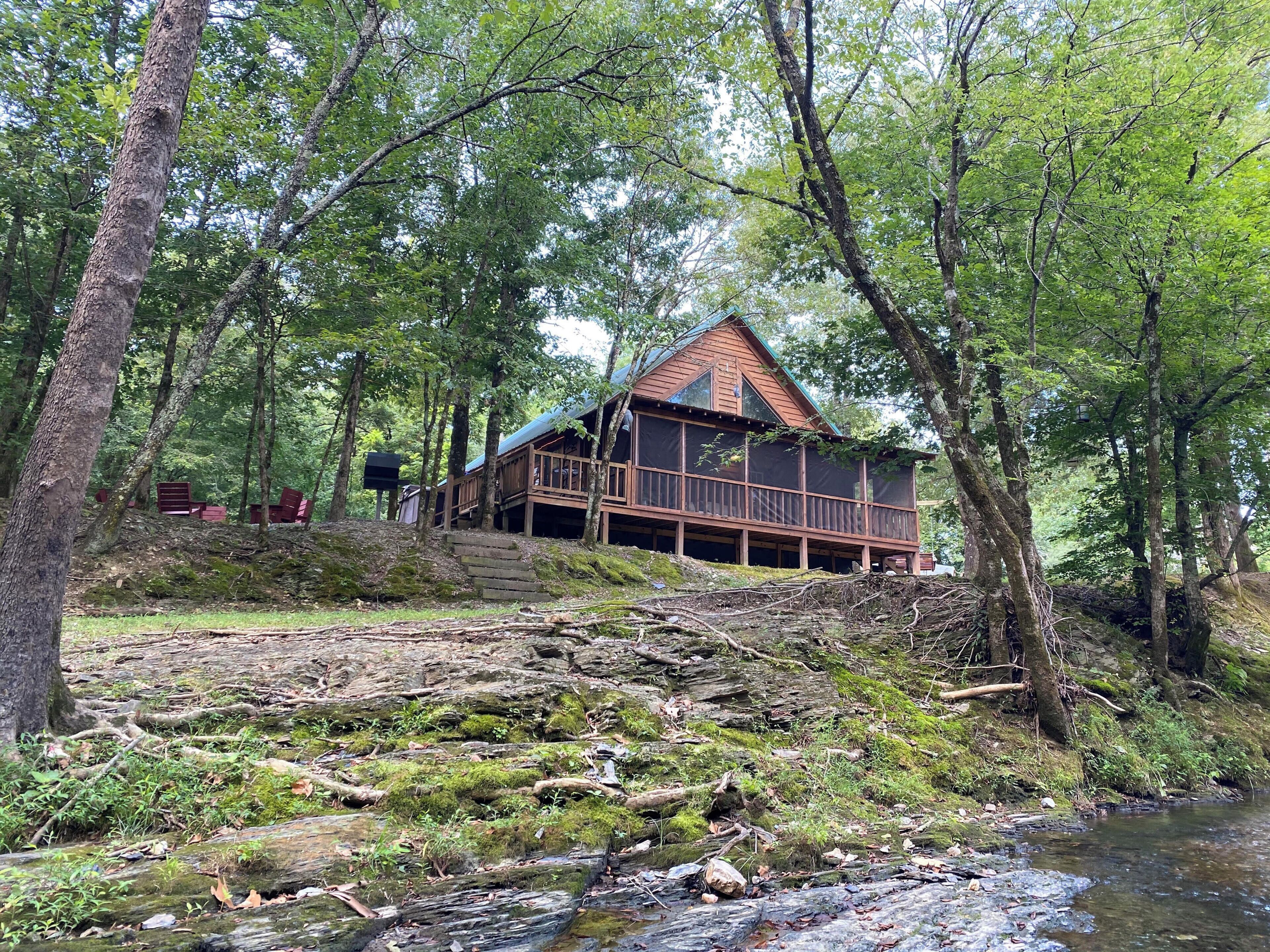 Une cabane dotée d’une terrasse, entourée d’arbres et bordée par un ruisseau.