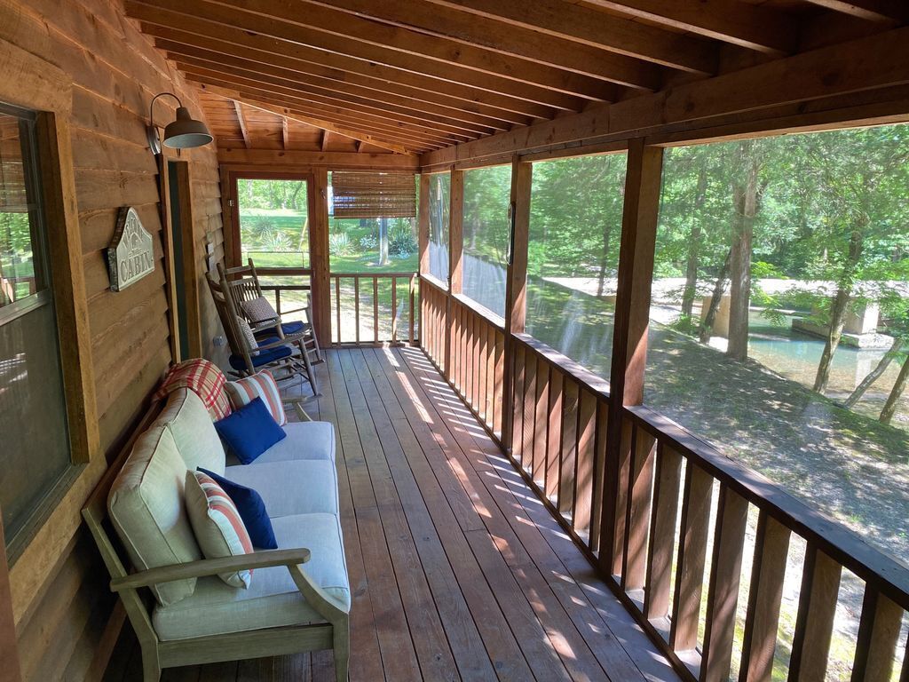 Eine überdachte Veranda mit einer Holzbank und Kissen, großen Fenstern und Blick auf Bäume.