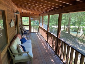 A covered porch with a wooden bench and cushions, large windows, and a view of trees.