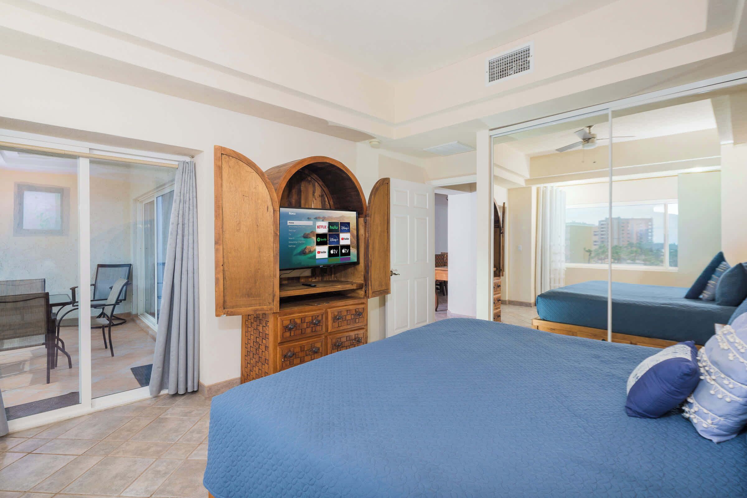 Ein Hotelzimmer mit einem großen Bett, einem an einer Holzkommode montierten Fernseher, einem Schreibtisch mit Stuhl und einer Schiebetür zu einem Balkon.