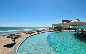 Blick über einen Pool mit Strand und Meer im Hintergrund, sowie ein Gebäude mit Kuppelbau.
