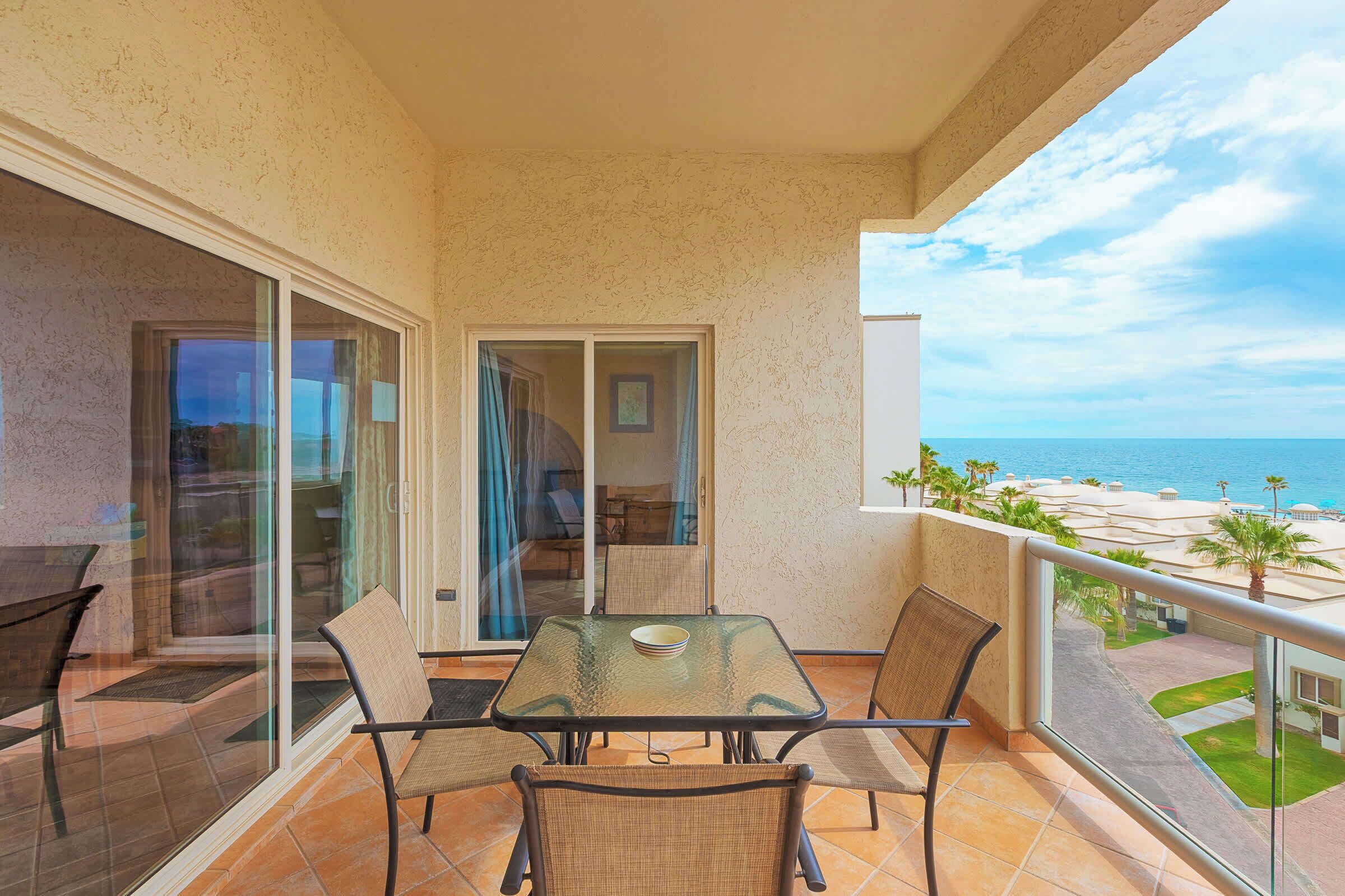 Ein Balkon mit Glastisch und Stühlen, Blick auf das Meer, und ein Gebäude mit einer Palme im Hintergrund.