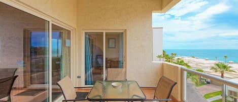 Ein Balkon mit Glastisch und Stühlen, Blick auf das Meer, und ein Gebäude mit einer Palme im Hintergrund.