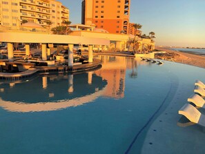 A pool with a curved edge, a lounge area with chairs, and a view of a beach and buildings in the background. - Las Palmas Delphin 402 Beachfront Paradise (Puerto Peñasco)