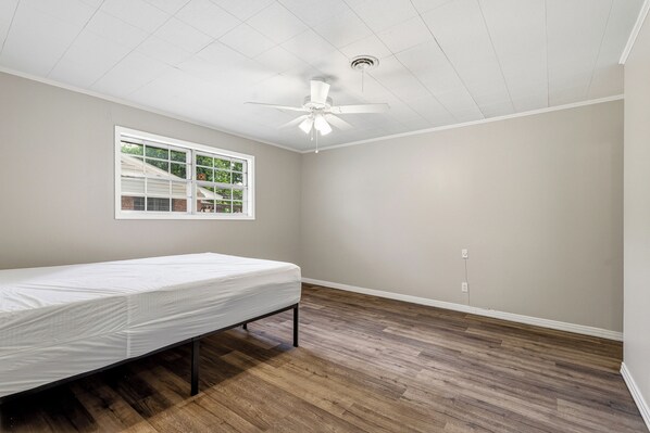 Une chambre avec un lit simple, un ventilateur de plafond, une fenêtre et un plancher en bois.