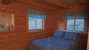 Une chambre à coucher dans une cabane en bois, avec un lit, deux fenêtres ornées de rideaux à carreaux, une lampe de chevet et un ventilateur.