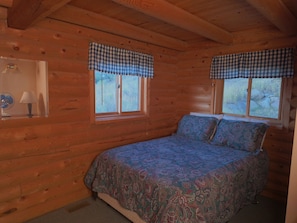 A wooden cabin bedroom with a bed, two windows with checkered valances, a bedside lamp, and a fan.