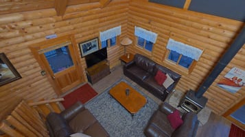 Interior de una cabaña de troncos con sofá, televisión, chimenea y ventana con cortina de encaje.