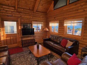 Interior de una cabaña de madera con televisión, sofás de cuero, una mesa de centro y ventanas con vistas.