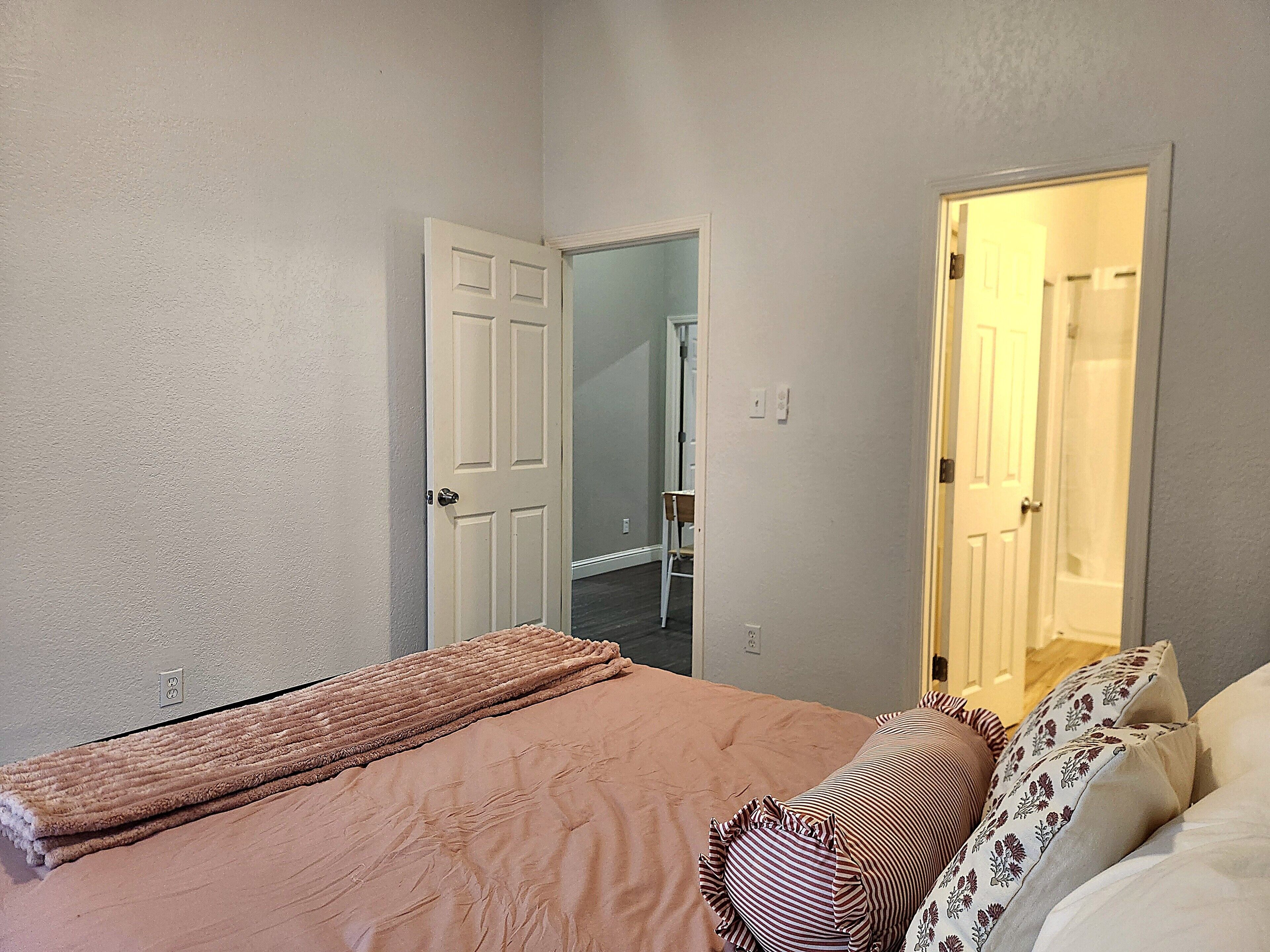 A bedroom with a bed, a door, and a visible bathroom in the background.