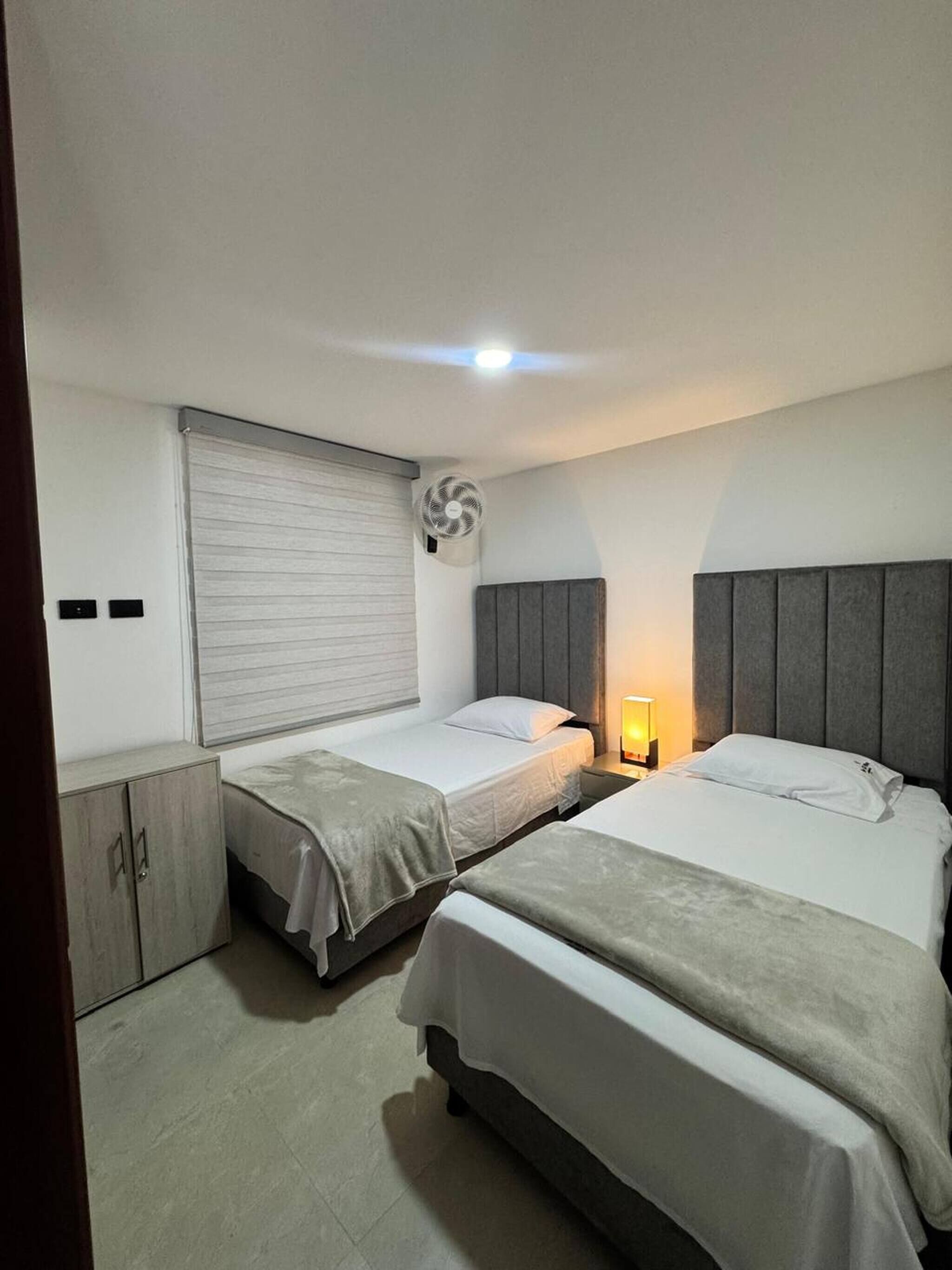 A hotel room with two beds, a wardrobe, a window with blinds, a standing fan, and a bedside lamp.