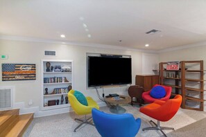 Ein modernes Wohnzimmer mit Flachbildfernseher, farbenfrohen Sesseln, einem Bücherregal und einem kleinen Couchtisch.