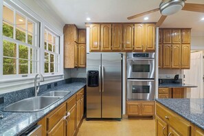 Cocina con refrigerador de acero inoxidable, dos hornos, cafetera integrada, gabinetes de madera, encimeras de granito y ventana con vista a árboles.
