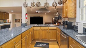 Une cuisine dotée de plans de travail en granit, de meubles en bois, d’une hotte de cuisinière en acier inoxydable, de casseroles suspendues et d’un paillasson immaculé.