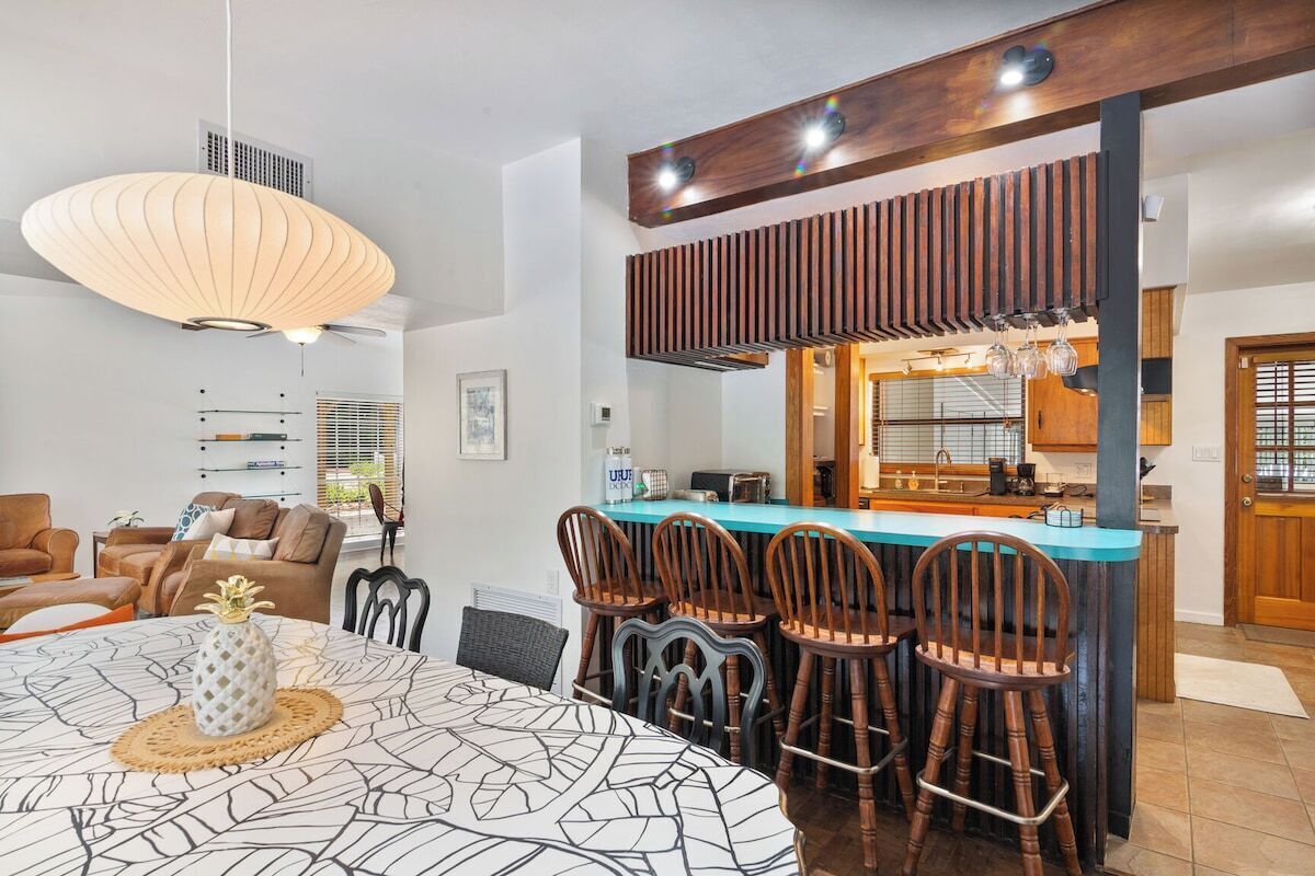 A modern kitchen with a central island, wooden chairs, and a dining table with a patterned tablecloth.