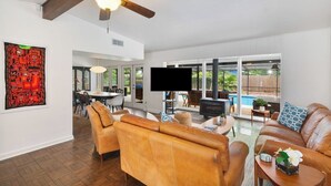 Un salón comedor amplio con ventilador de techo, televisor de pantalla plana, sofás de cuero y un gran ventanal con vistas a un patio.