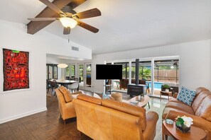 A spacious living room with a ceiling fan, a flat-screen TV, leather sofas, and a large window overlooking a backyard. - Gator Pool Home - Close to UF (Gainesville)
