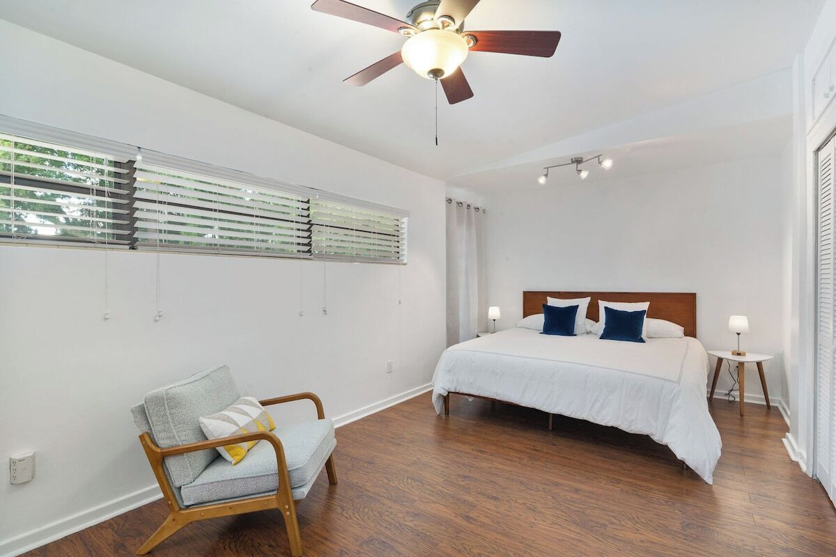 Un dormitorio con cama, una silla, ventilador de techo y ventana con persianas.