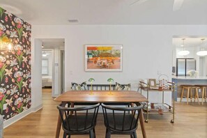 Un comedor con mesa y sillas de madera, un cuadro en la pared y una cocina al fondo.