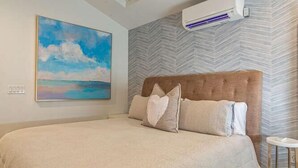 Ein Schlafzimmer mit einem großen Bett, einem Kopfteil, einer wandmontierten Klimaanlage und einem Gemälde mit Strandmotiv.